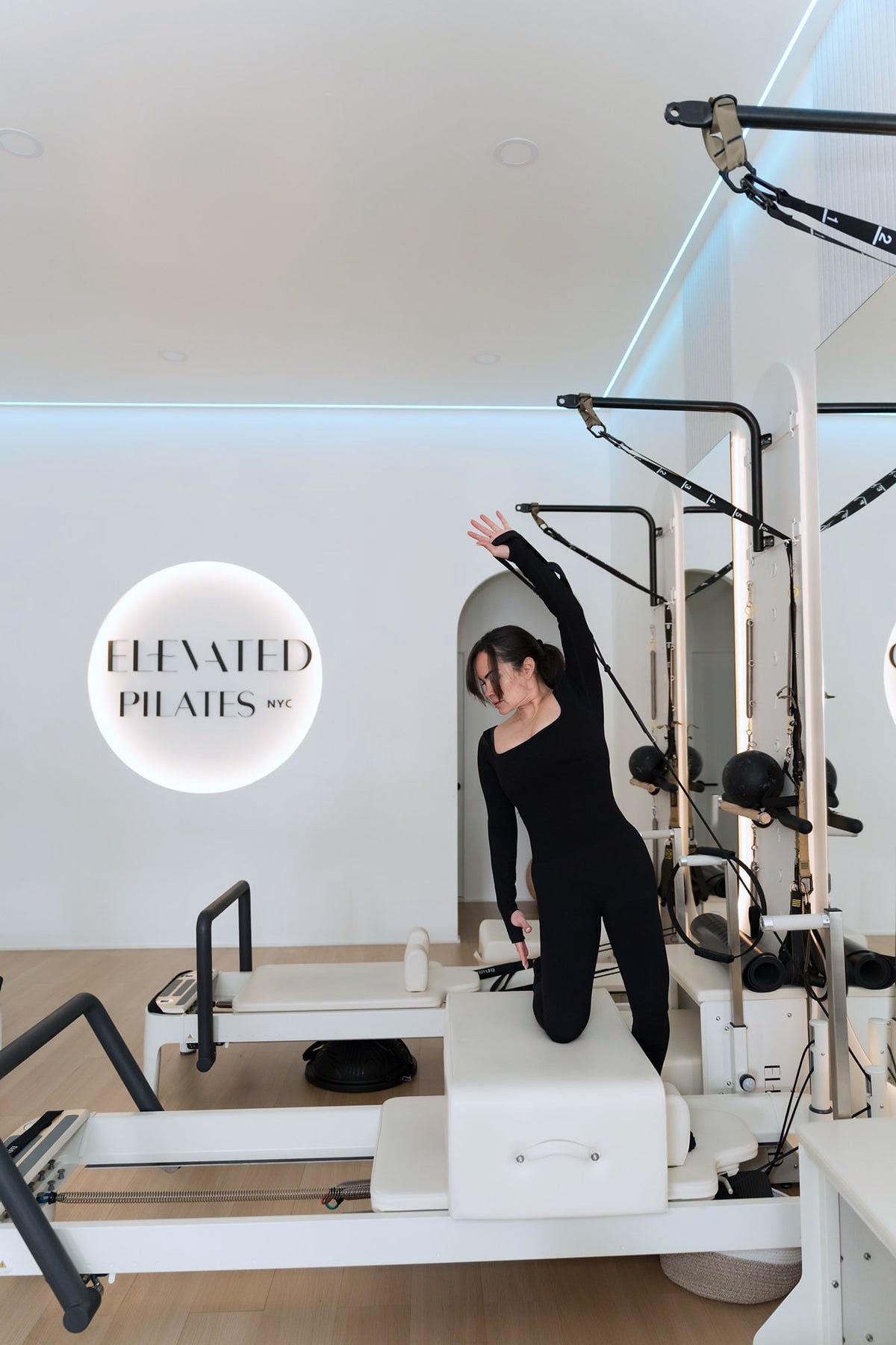 Woman kneeling on reformer reaching up in studio with lit logo sign