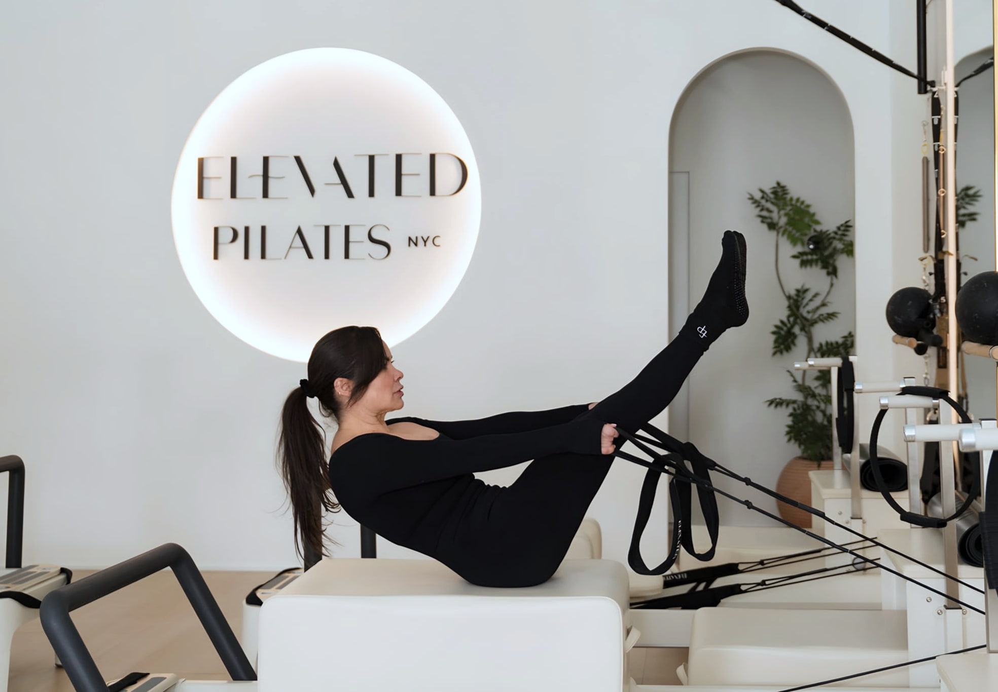 Woman on reformer with legs raised beneath illuminated Elevated Pilates NYC sign